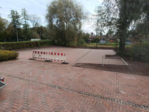 Neu gepflasterte Fläche mit roten Klinkersteinen, umgeben von Hecken und Bäumen, eine mobile Absperrung steht im Vordergrund.