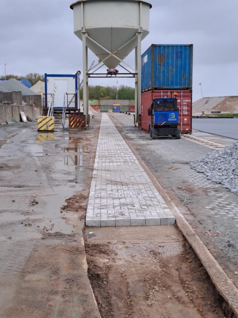 Frisch verlegter Gehweg aus Pflastersteinen entlang eines Industriehafens, mit Container, Maschinen und Lagerfläche im Hintergrund.
