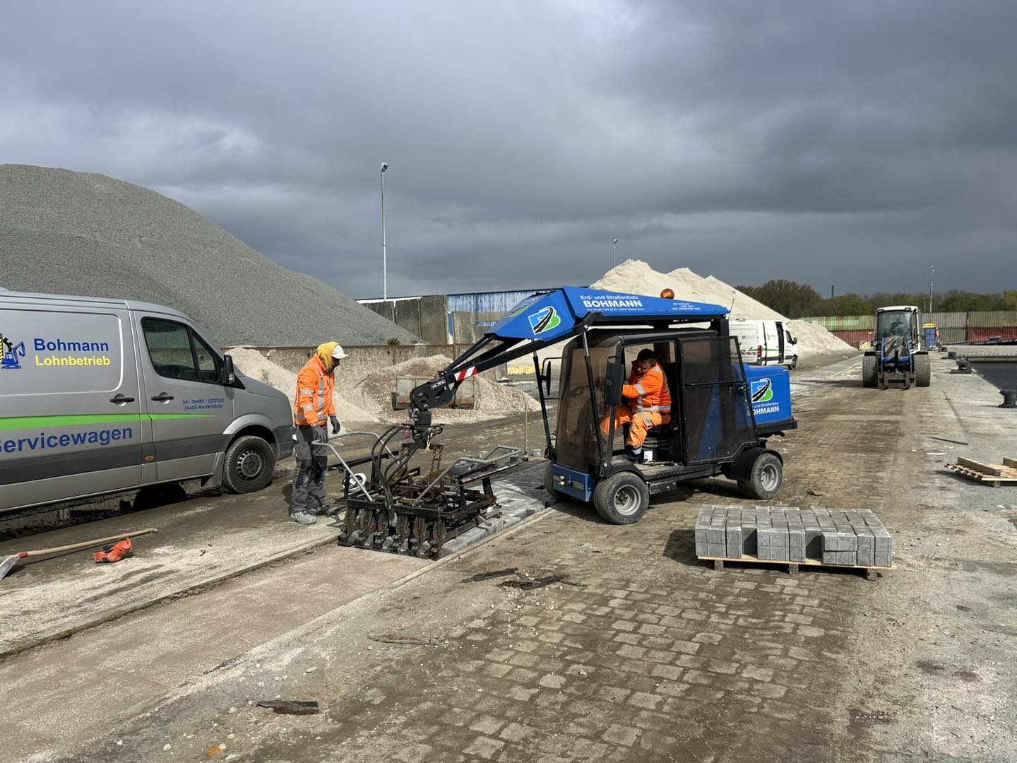 Zwei Arbeiter verlegen mit maschineller Unterstützung Pflastersteine auf einem Industriegelände, Servicefahrzeug und Material im Hintergrund.