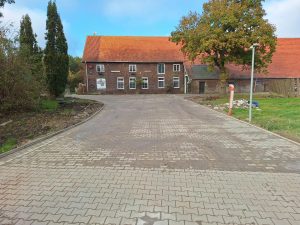 Gepflasterte Hoffläche aus Beton-Verbundpflaster vor einem historischen Ziegelgebäude mit rotem Dach und großem Baum.