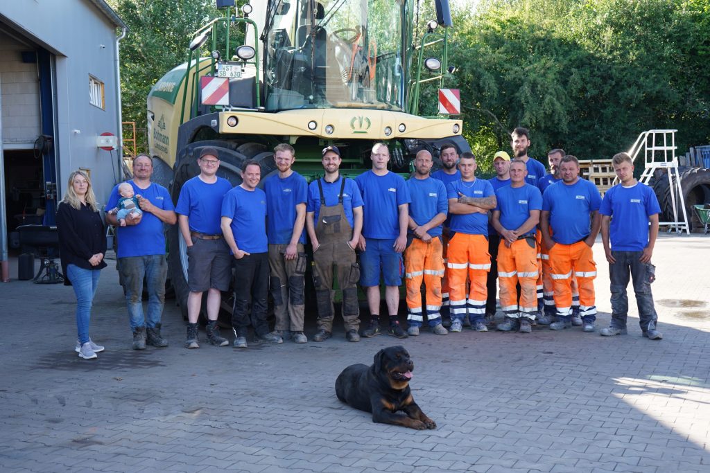 Gruppenfoto des Teams von Erd- und Straßenbau Bohmann mit einem Hund im Vordergrund, aufgenommen auf dem Betriebshof vor einer großen Arbeitsmaschine.