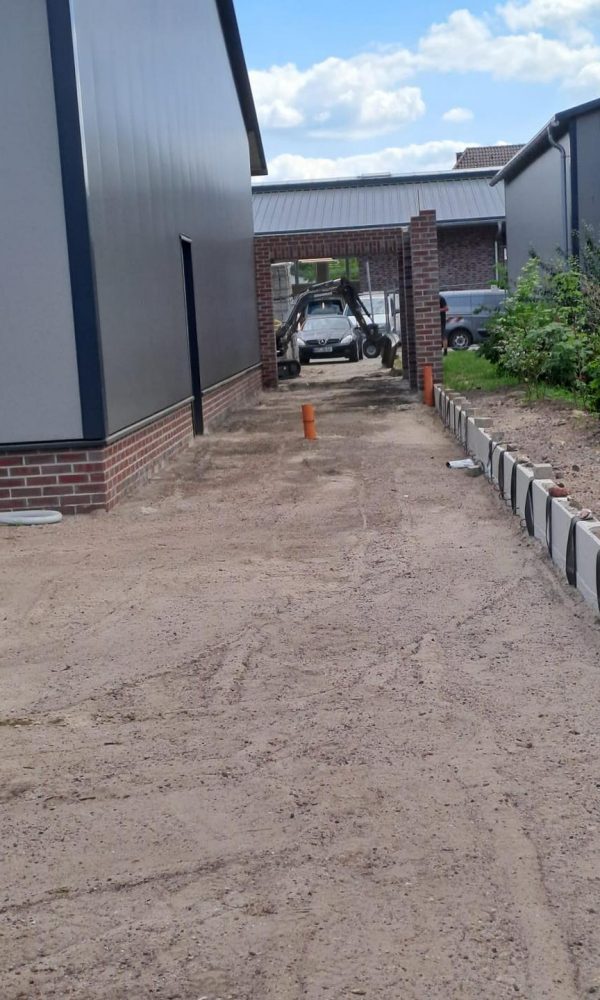 Verdichteter Sanduntergrund zwischen zwei Gebäuden mit seitlicher Bordsteineinfassung, im Hintergrund ein Carport und geparkte Fahrzeuge.