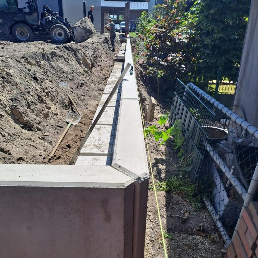 Lange Winkelstützwand aus Beton zur Hangsicherung zwischen zwei Grundstücken, mit Baustelle, Bagger und gespannter Schnur zur Ausrichtung.