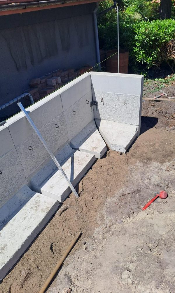 Im Bau befindliche Winkelstützwand aus Betonfertigteilen, aufgestellt an einer Hauswand auf verdichtetem Erdreich.