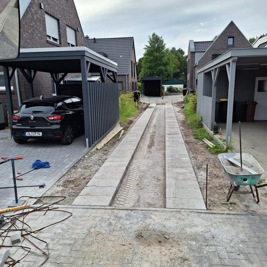 Baustelle einer neuen Zufahrt mit Fahrspuren aus Betonplatten zwischen zwei modernen Wohngebäuden mit Carports.
