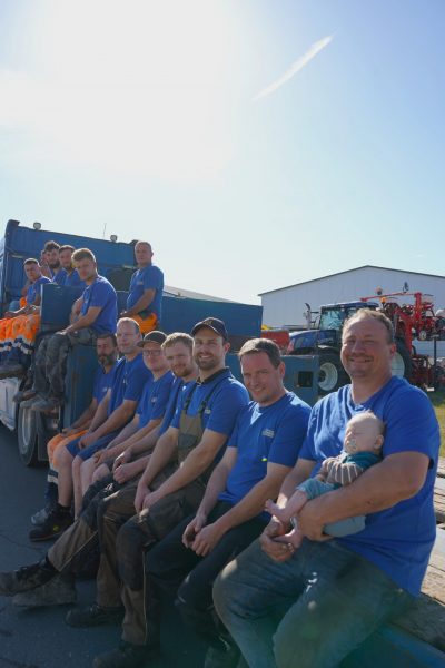 Gruppenfoto des Teams von Erd- und Straßenbau Bohmann, aufgenommen im Freien bei Sonnenschein. Die Männer tragen Arbeitskleidung, lächeln in die Kamera, ein Kind wird von einem Teammitglied auf dem Schoß gehalten.