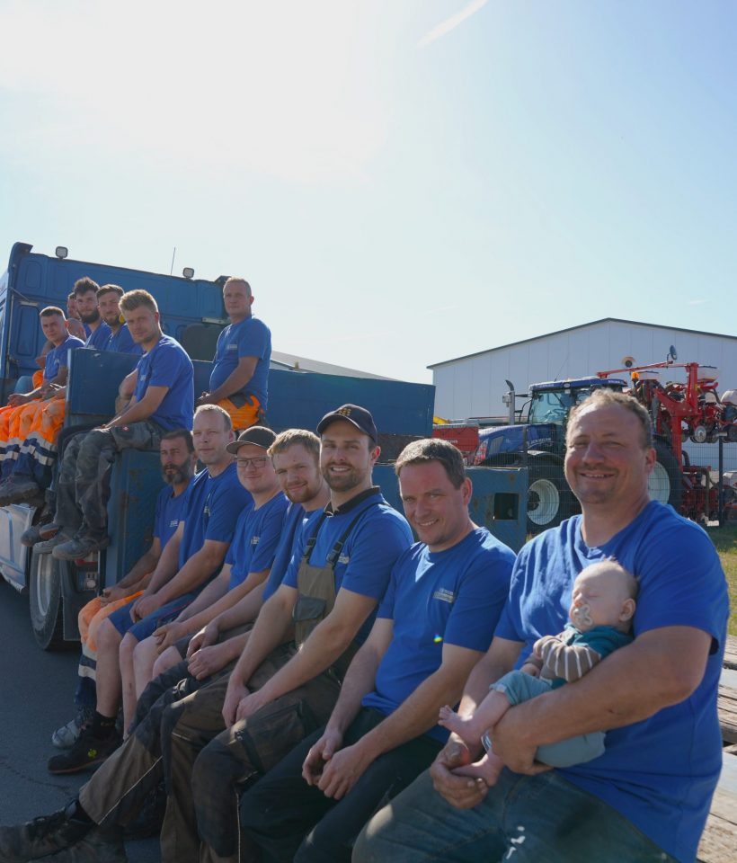 Gruppenfoto des Teams von Erd- und Straßenbau Bohmann, aufgenommen im Freien bei Sonnenschein. Die Männer tragen Arbeitskleidung, lächeln in die Kamera, ein Kind wird von einem Teammitglied auf dem Schoß gehalten.